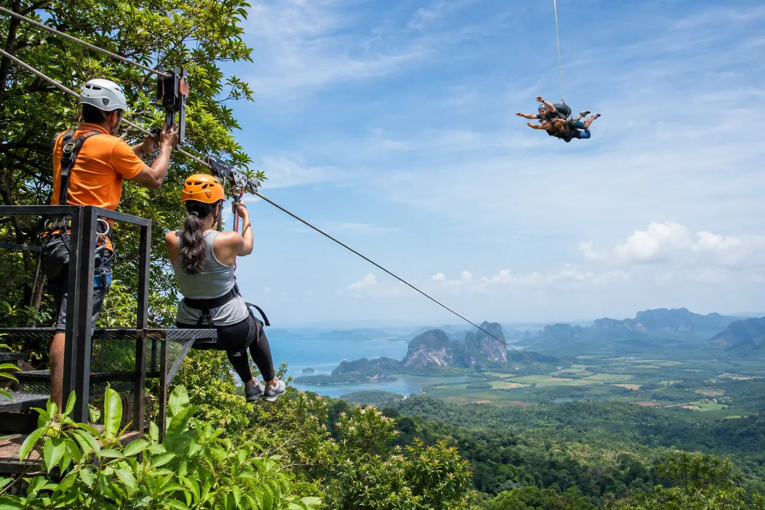 A tandem skydiving pair in freefall above a coastal landscape in southern Thailand, with an instructor preparing a second jump from a platform.