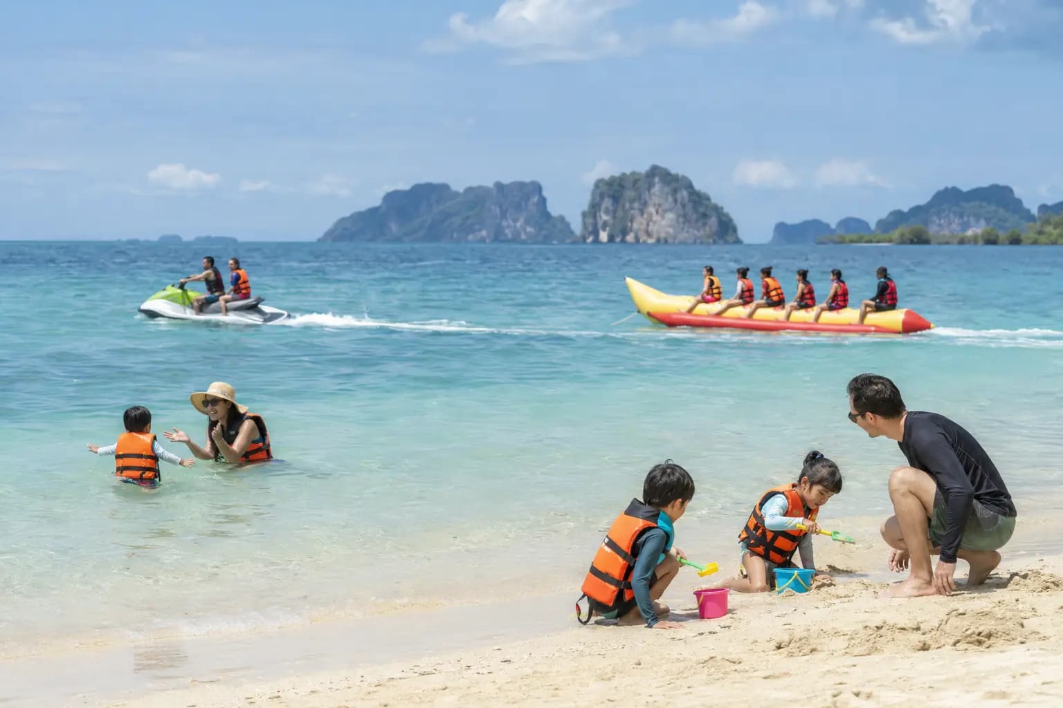 A family playing safely on the sand in life jackets while a jet ski and banana boat operate further out at sea in Thailand.